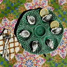 A green ceramic oyster shell platter with six indentations, styled with six raw oysters, two lemon wedges, forks, and a napkin on a vibrant floral tablecloth.