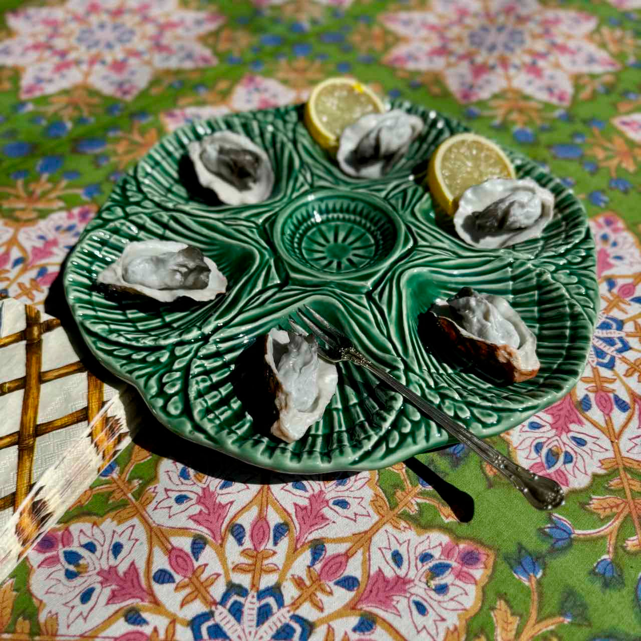 A green ceramic octagonal serving platter holding six raw oysters on a patterned tablecloth, with two lemon wedges and a small spoon.