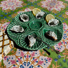 A green ceramic octagonal serving platter holding six raw oysters on a patterned tablecloth, with two lemon wedges and a small spoon.