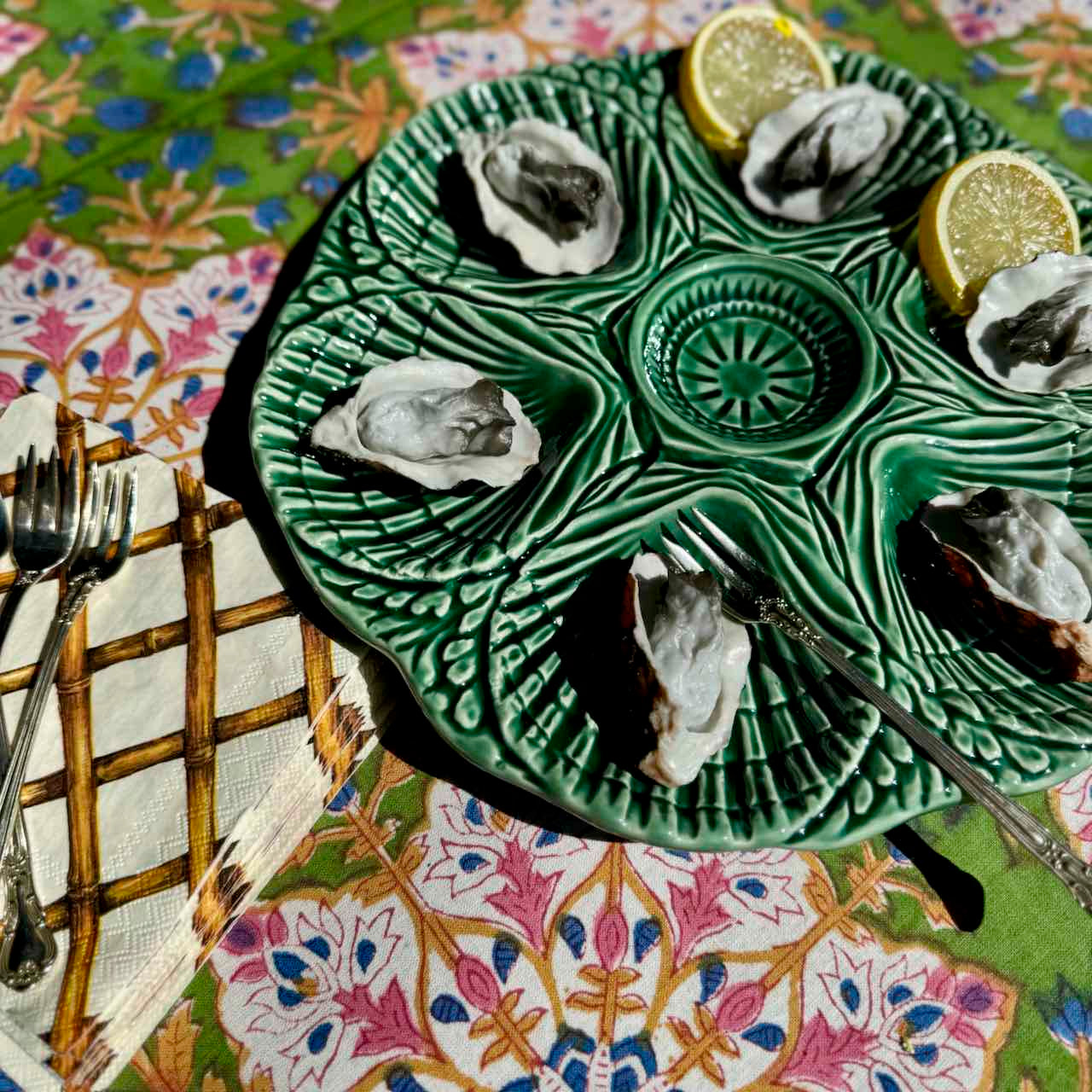 A green ceramic oyster platter with six compartments, filled with raw oysters and garnished with lemon slices, set on a colorful patterned tablecloth next to silver forks.