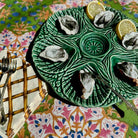 A green ceramic oyster platter with six compartments, filled with raw oysters and garnished with lemon slices, set on a colorful patterned tablecloth next to silver forks.