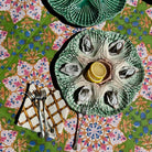 A top-down view of a green ceramic oyster shell platter holding several oysters and a lemon slice, set on a colorful floral tablecloth with cutlery nearby.