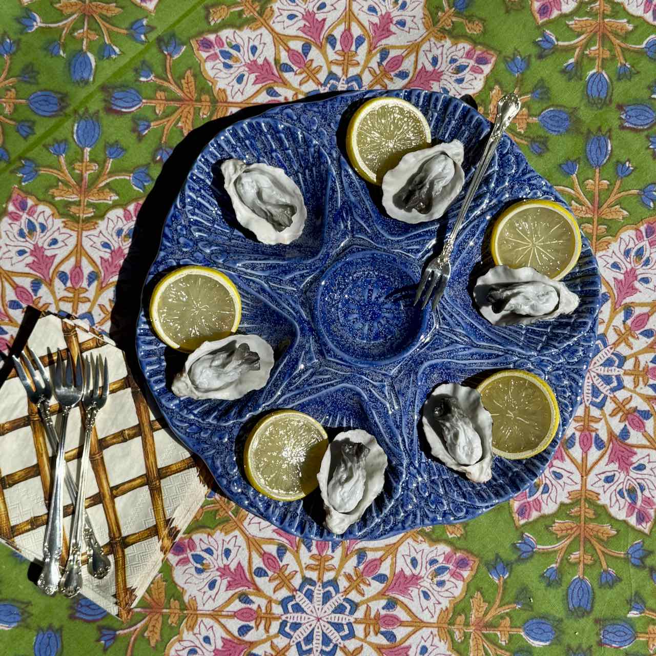 A top-down view of a blue ceramic oyster serving platter with eight compartments, filled with raw oysters and lemon slices, set on a colorful patterned tablecloth with silver forks and a napkin.