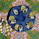A top-down view of a blue ceramic oyster serving platter with eight compartments, filled with raw oysters and lemon slices, set on a colorful patterned tablecloth with silver forks and a napkin.