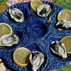 A blue ceramic octagonal serving platter holding six raw oysters on a bed of white sauce, garnished with lemon slices, with a metal serving fork resting on top.