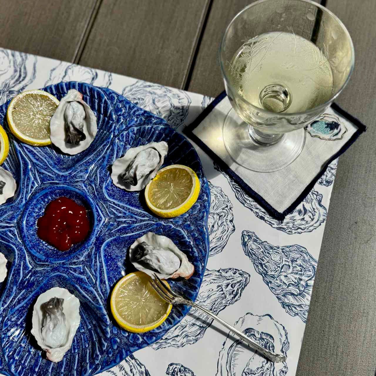 A blue ceramic platter with shell-shaped cutouts holds several oysters, lemon slices, a small bowl of red sauce, and a fork, arranged on a white placemat with blue oyster illustrations. A glass of white wine and a napkin are also visible on a gray wooden table.