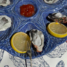 A blue ceramic platter holding several oysters on the half, with lemon wedges and a small bowl of red sauce, photographed close up on a patterned table surface.