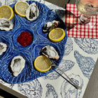 A blue ceramic oyster platter with lemon slices and a small red dipping sauce, set on a patterned tablecloth next to a red-and-white checkered napkin and a wine glass.