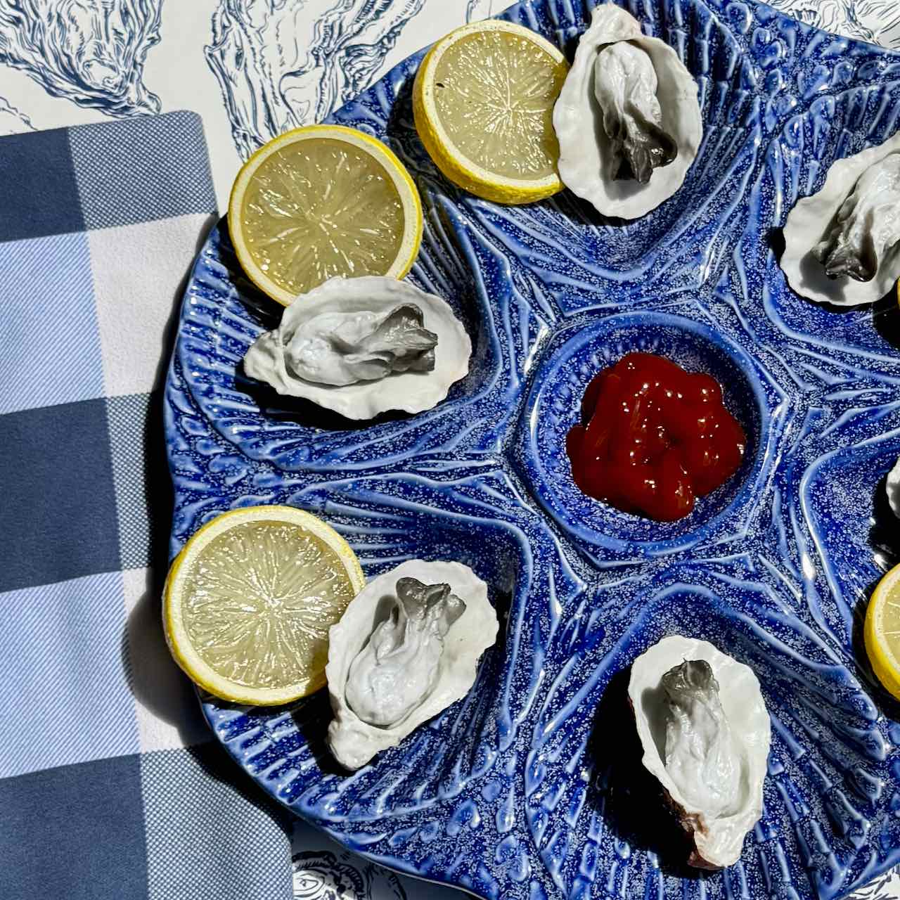 A blue ceramic platter with a textured leaf-like pattern, arranged with raw oysters, lemon slices, and a small red dipping sauce in the center, set on a blue-and-white checkered napkin.