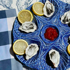 A blue ceramic platter with a textured leaf-like pattern, arranged with raw oysters, lemon slices, and a small red dipping sauce in the center, set on a blue-and-white checkered napkin.