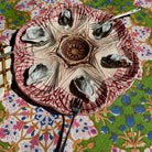 A decorative ceramic oyster shell dish with six indentations around a central well, holding several raw oysters, set on a colorful floral-patterned tablecloth with forks placed around it.