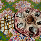 A decorative ceramic oyster shell platter with six compartments, shown on a colorful floral tablecloth with forks and a napkin beside it.