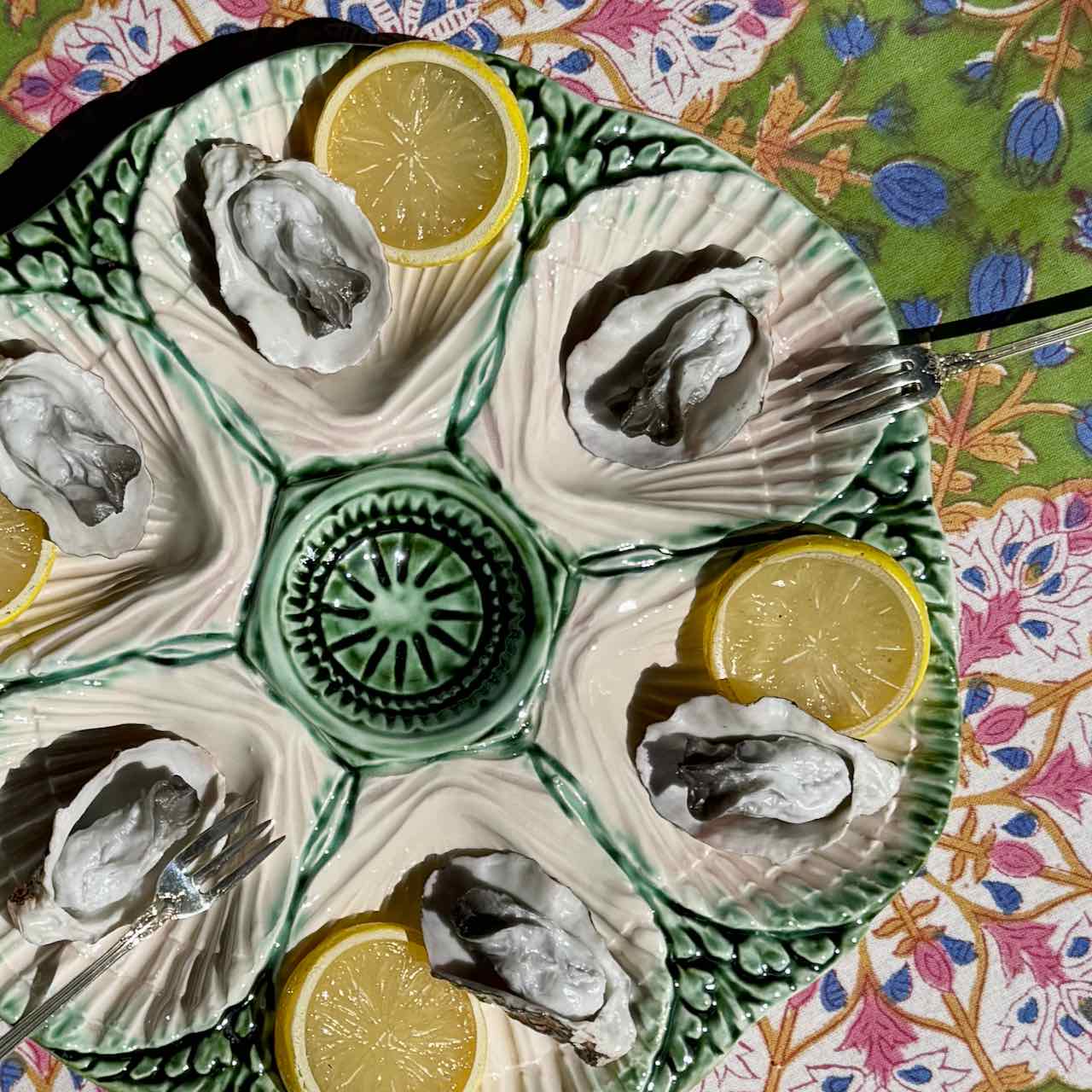 A ceramic oyster shell platter with six compartments, each holding an open oyster and a lemon slice, set on a colorful floral-patterned tablecloth.