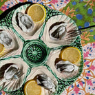 A ceramic oyster shell platter with six compartments, each holding an open oyster and a lemon slice, set on a colorful floral-patterned tablecloth.