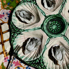 A ceramic oyster shell platter with four compartments and a central dipping bowl, shown filled with raw oysters. The platter has a white and green shell-like design and is photographed on a patterned tablecloth with a fork visible at the lower left.