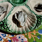 A close-up of a ceramic oyster shell serving dish holding several raw oysters, set on a colorful floral-patterned fabric surface.