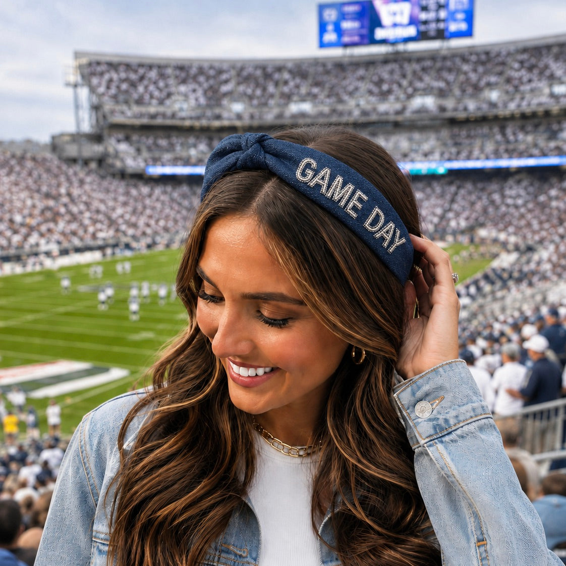 A woman in a stadium wearing a navy blue denim headband with 'GAME DAY' embroidered in white, paired with a light wash denim jacket over a white top.
