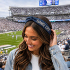 A woman in a stadium wearing a navy blue denim headband with 'GAME DAY' embroidered in white, paired with a light wash denim jacket over a white top.