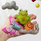 Hand holding felt animal toys including a frog, butterfly, and snail against a white background.
