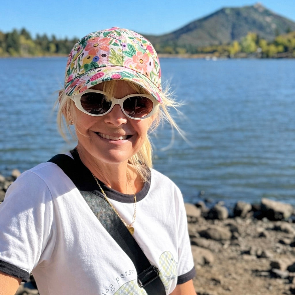 Woman wearing a floral cap and sunglasses by a lake with mountains in the background