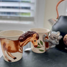 Mug with horse design, another mug with koala design, and a teapot on a surface.