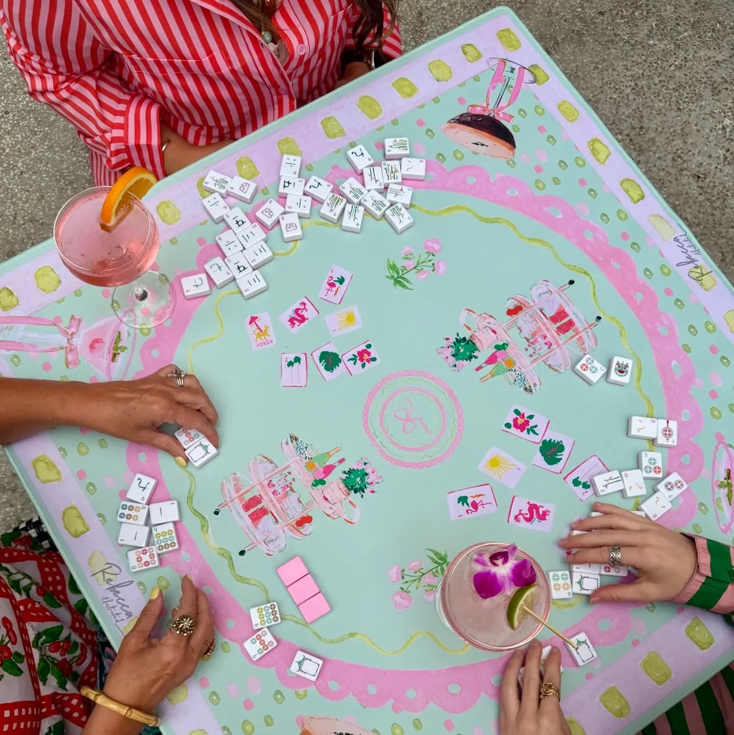 An overhead view of a colorful Mahjong game board with multiple players' hands visible placing tiles. The board features a pastel pink and mint green design with cartoon illustrations of a bunny, a woman, and flowers.