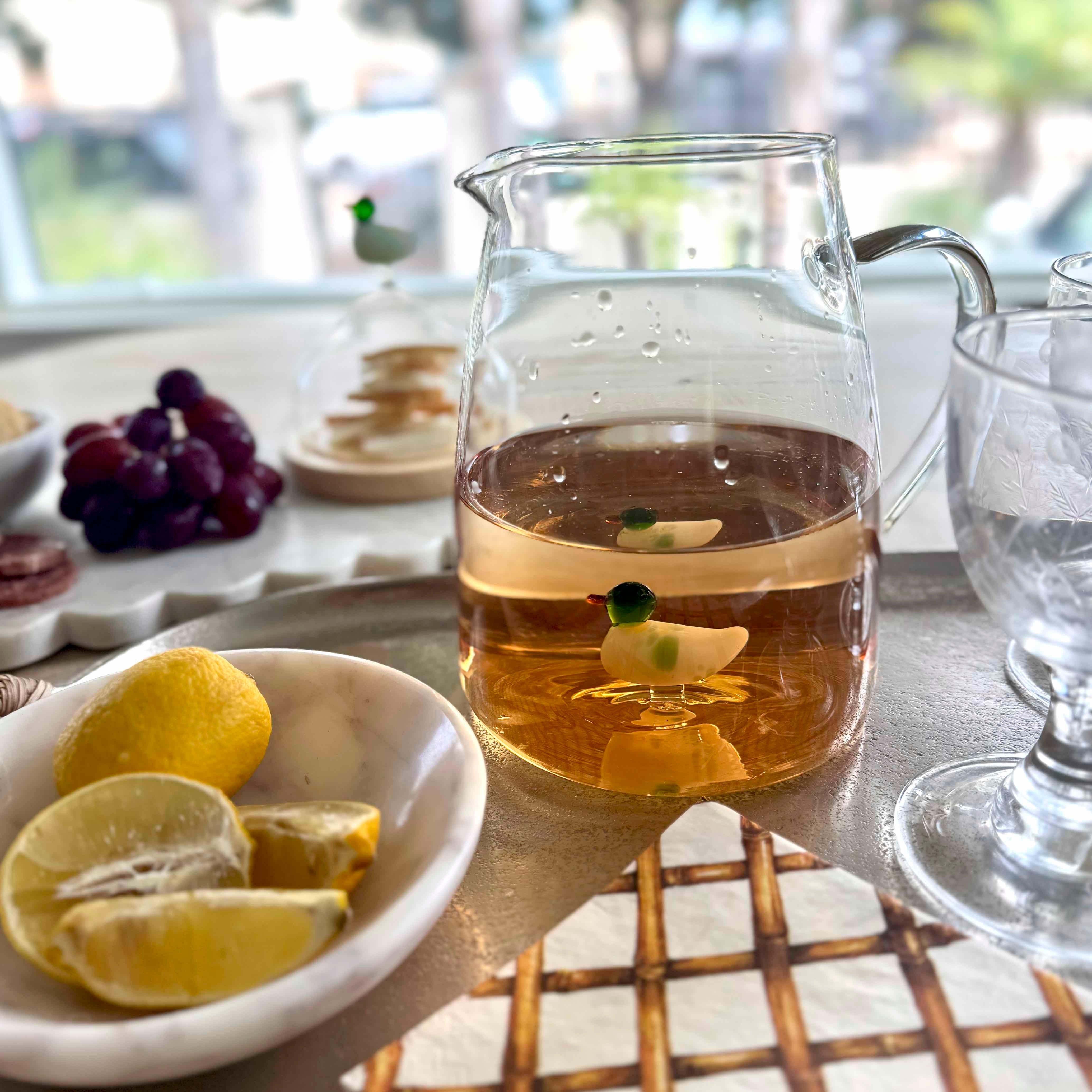 Glass pitcher of tea with lemon slices and a duck toy on a table with lemons and grapes.