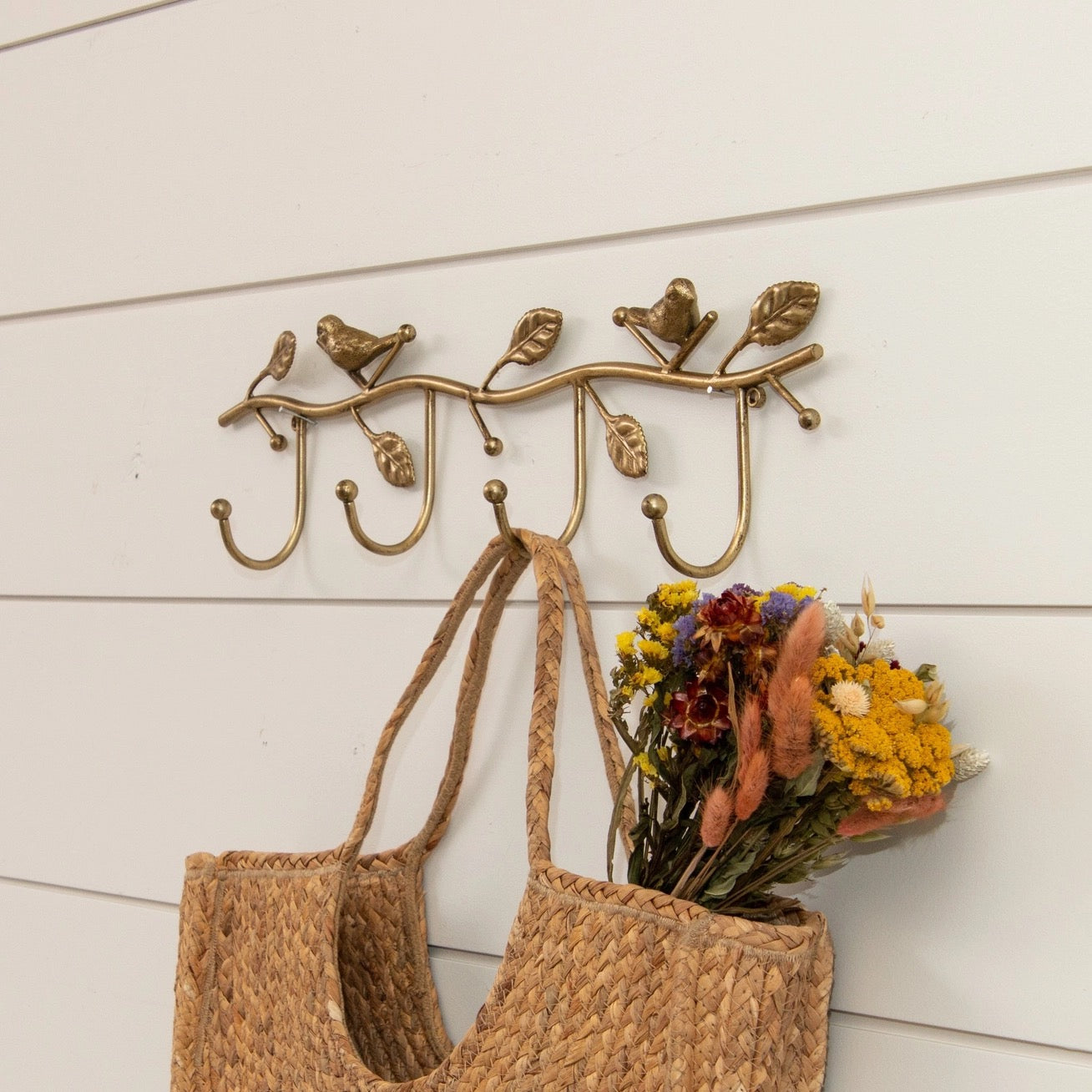 Decorative wall hook with birds and leaves design, holding a woven bag and flowers against a white wooden background.