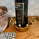 A gold crown-shaped wine bottle holder cradling a dark wine bottle on a wooden tray, with a glass and a checkered napkin nearby.