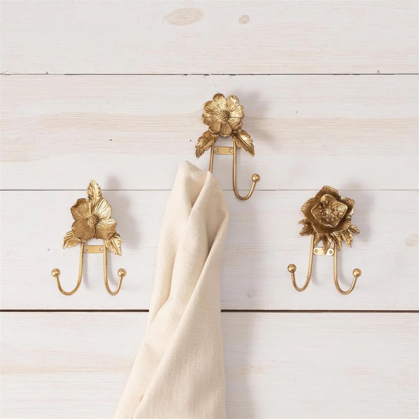 Three gold floral-patterned hooks on a wooden wall with a beige towel draped over one of them.