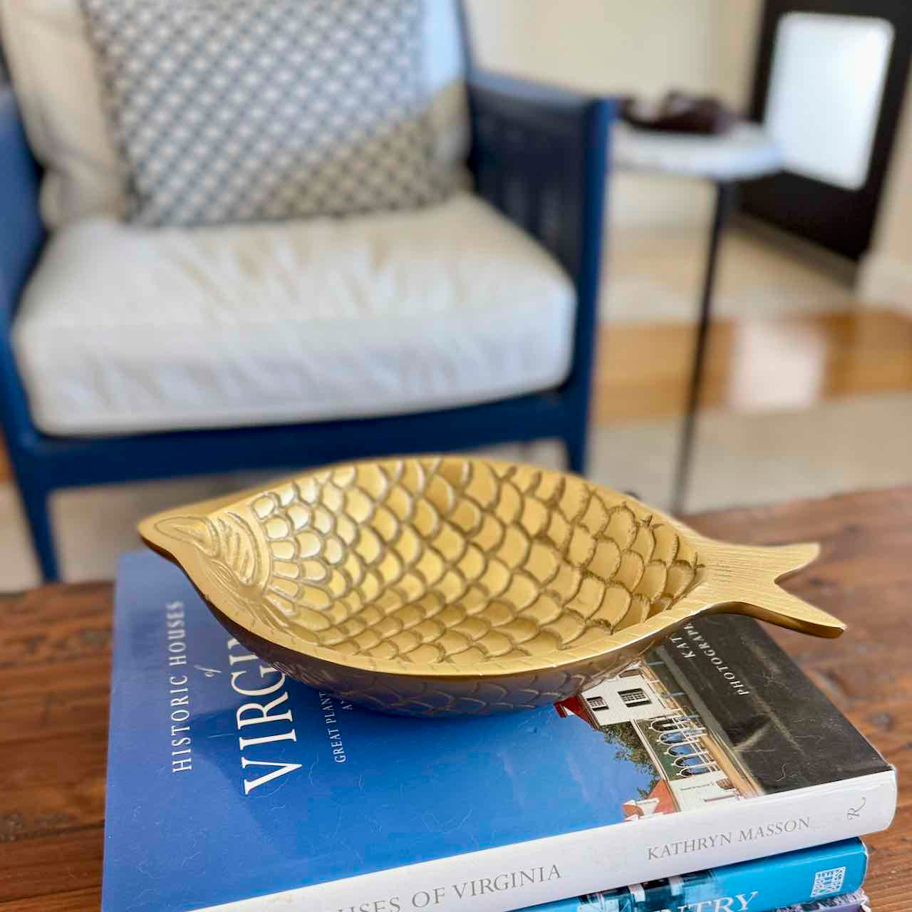 A gold fish-shaped decorative bowl with a textured scale pattern, resting on a stack of books on a wooden table in a living room setting.