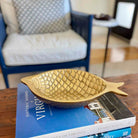 A gold fish-shaped decorative bowl with a textured scale pattern, resting on a stack of books on a wooden table in a living room setting.