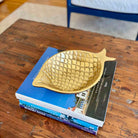 A gold fish-shaped decorative tray with a textured, scale-like pattern, resting on a stack of books on a rustic wooden table.