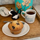 A breakfast scene on a wooden tray with a blueberry muffin on a white plate, a white mug with blue script text, a small black-and-white cow-shaped pitcher, and a turquoise decorative container in the background.