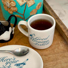 A white ceramic mug filled with tea sits on a wooden surface, printed with the phrase 'Good Morning Ashley' in blue script. A matching white plate with a spoon is partially visible in the foreground, and a turquoise decorative container appears in the background.
