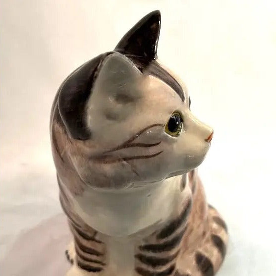 A ceramic cat figurine in a sitting position, featuring a cream-colored body with dark brown tabby stripes and a black-tipped ear.