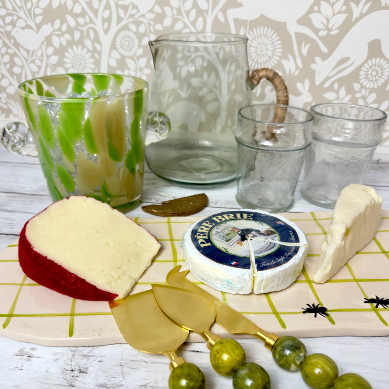 Cheese platter with various types of cheese, a knife set, and glassware on a decorative surface.