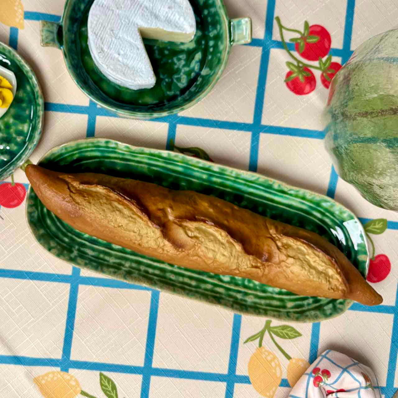 A long baguette rests on a green striped ceramic serving platter placed on a patterned tablecloth. Surrounding the platter are matching green ceramic dishes and food props, including a slice of cheese, a lemon, and a pear, creating a styled tabletop scene.