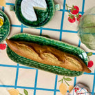 A long baguette rests on a green striped ceramic serving platter placed on a patterned tablecloth. Surrounding the platter are matching green ceramic dishes and food props, including a slice of cheese, a lemon, and a pear, creating a styled tabletop scene.