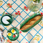 A styled overhead flat-lay of ceramic tableware on a printed tablecloth. The set includes a green speckled mug with a slice of white cake, a green oval platter holding a loaf of bread, a green round plate with halved hard-boiled eggs and small red and yellow accents, and a small green dish with a striped handle.