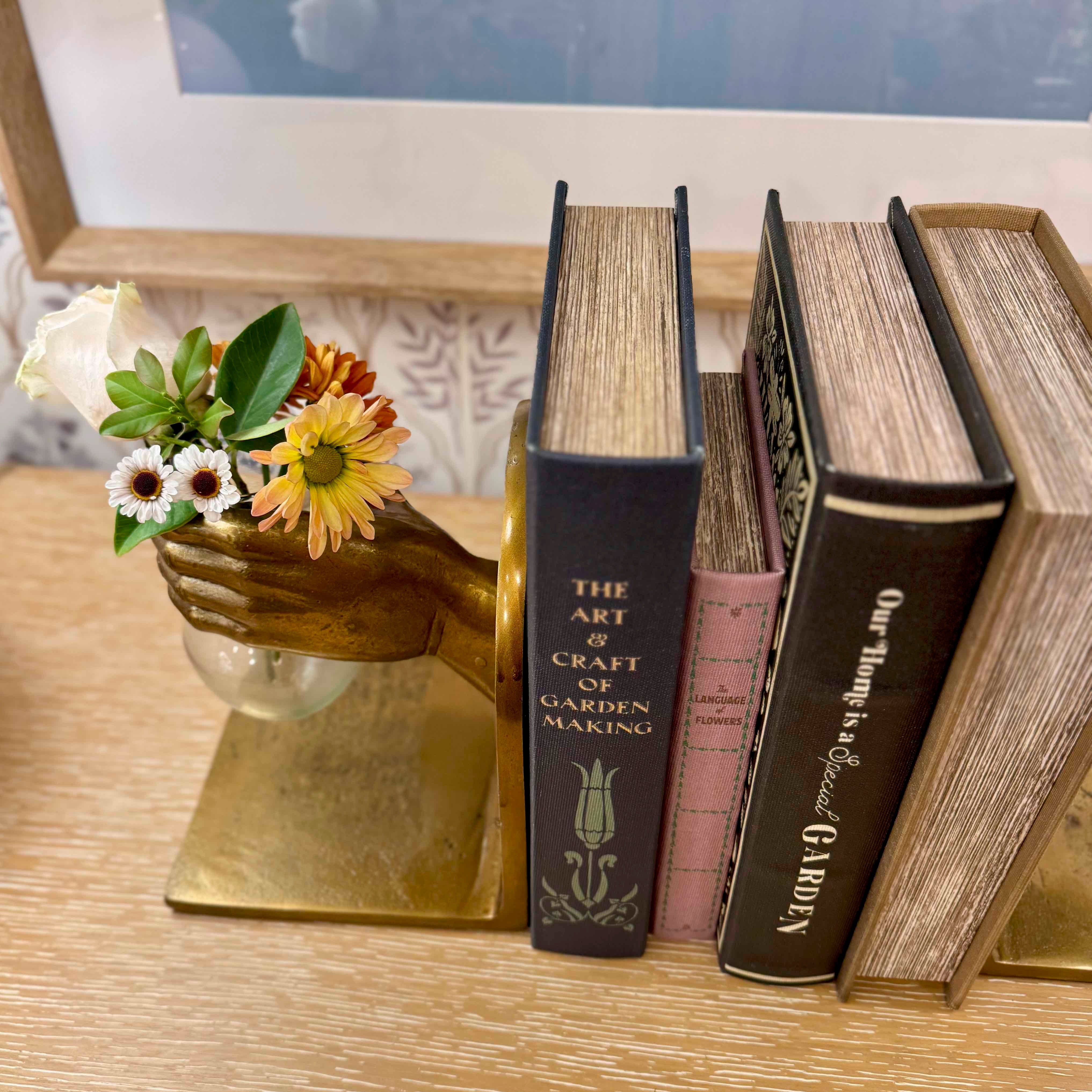 Decorative setup with books, a brass hand holding flowers, and a framed picture on a wooden surface.