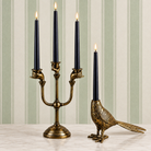 Two ornate brass candle holders on a light marble surface against a striped wallpaper background. One holder features three curved arms held by hands and holds three black taper candles lit at the top.