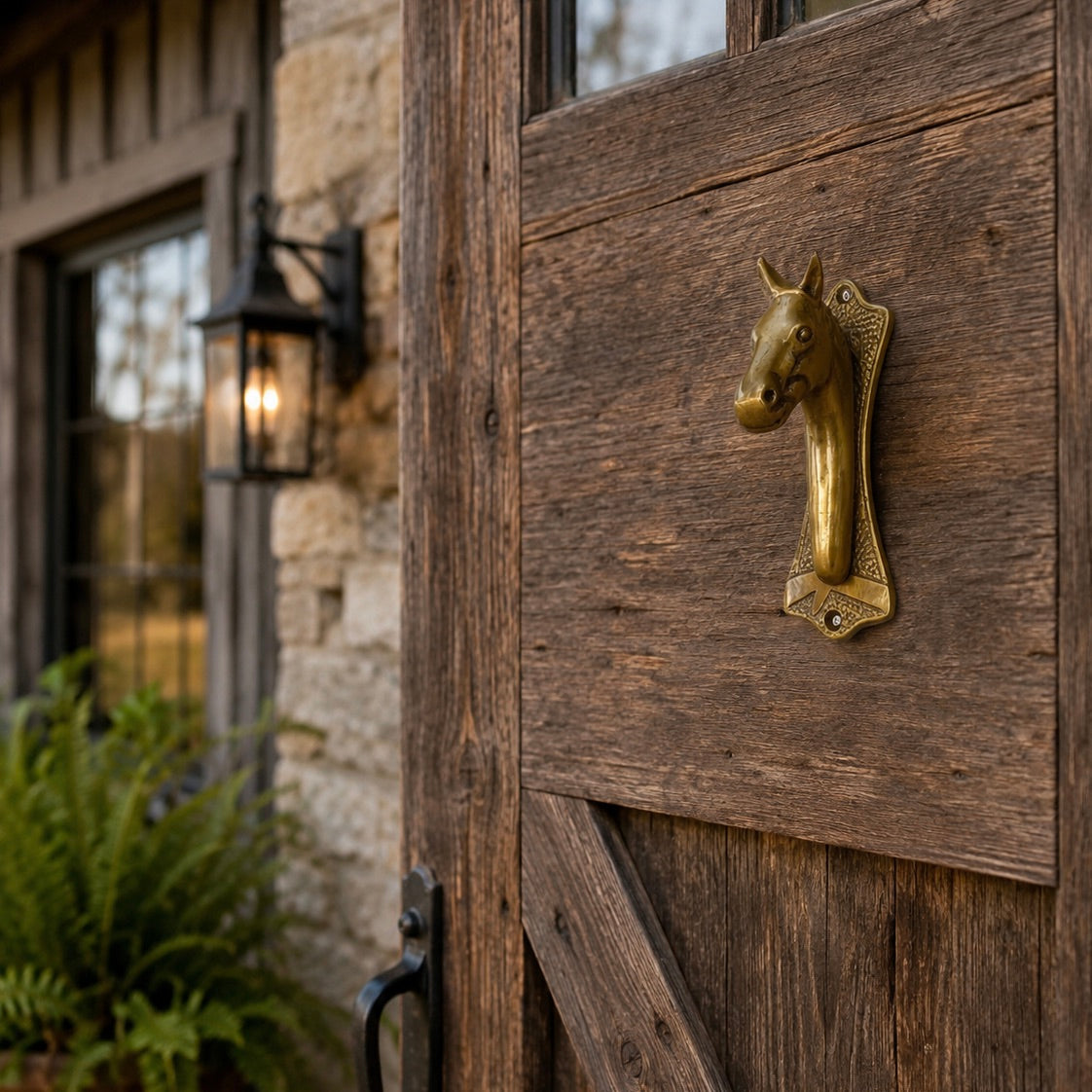 A rustic wooden barn-style door with a decorative brass horse-head door knocker mounted on the right side, shown outdoors next to a stone wall and a lit wall lantern.