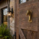 A rustic wooden barn-style door with a decorative brass horse-head door knocker mounted on the right side, shown outdoors next to a stone wall and a lit wall lantern.