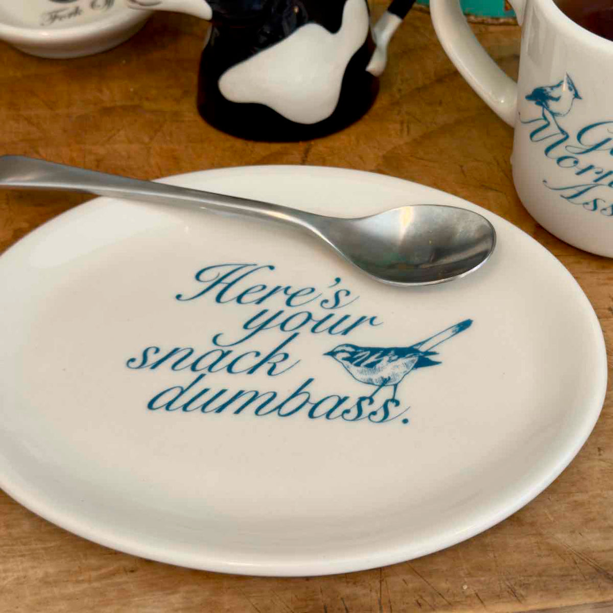 A white ceramic plate with blue text reading 'Here's your snack dumbass.' and a blue bird illustration, shown on a wooden table with a metal spoon resting across the top edge.