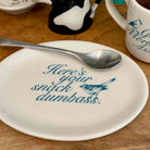 A white ceramic plate with blue text reading 'Here's your snack dumbass.' and a blue bird illustration, shown on a wooden table with a metal spoon resting across the top edge.