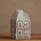 A decorative house-shaped paper lantern or box with a white base, hand-drawn black line art, and red floral and animal motifs, standing on a wooden surface against a textured beige wall.