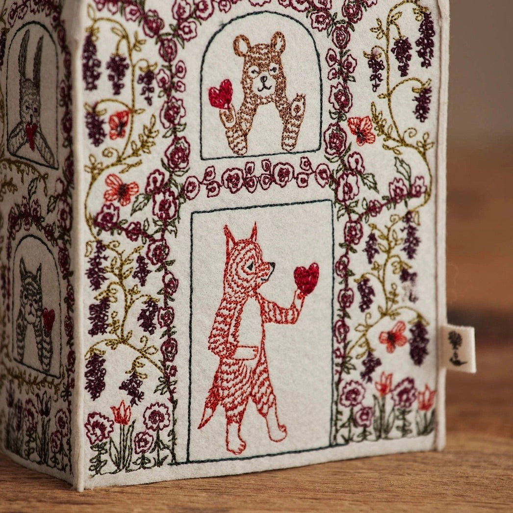 A close-up of a decorative fabric box or candle holder with embroidered animal and floral designs, shown on a wooden surface. The white fabric features stitched scenes of a fox, a bear, and a rabbit surrounded by flowers and vines, with red hearts held by the animals.