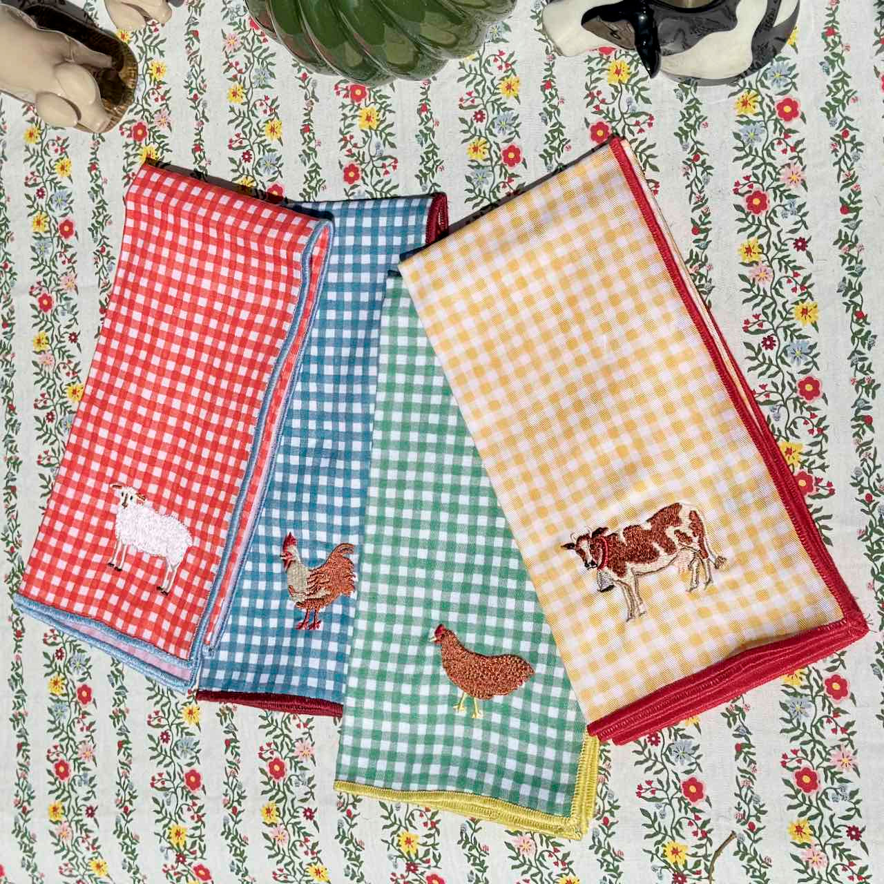 Set of four checkered towels with embroidered animals on a floral-patterned tablecloth.