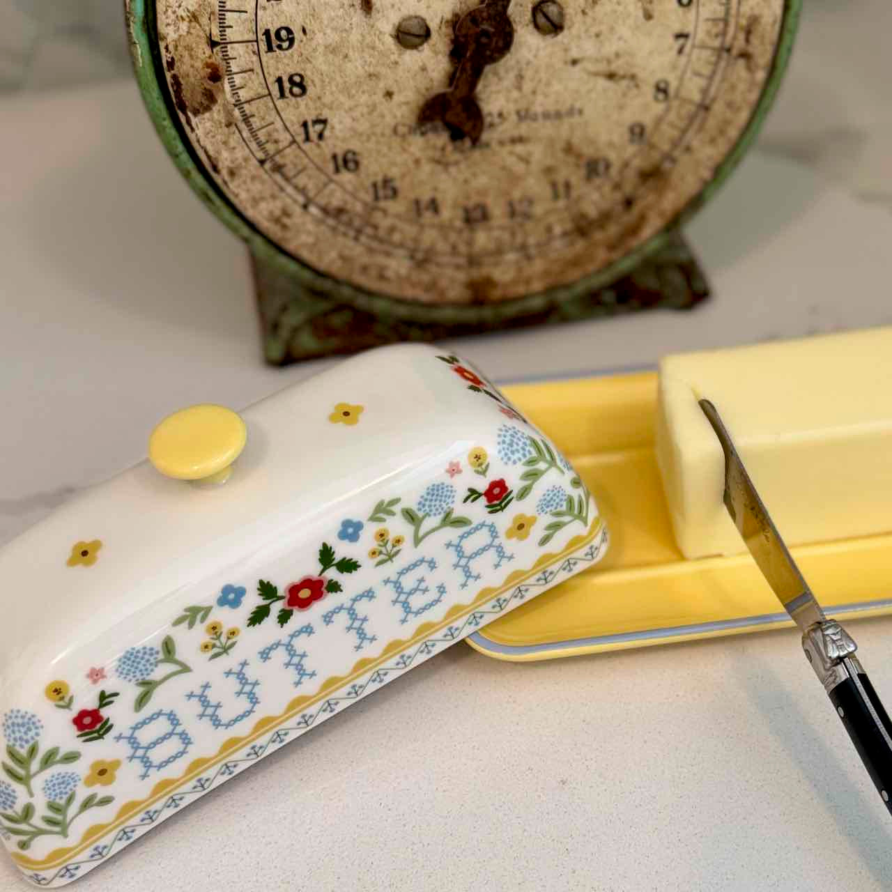 Butter dish with floral design, block of butter, and knife on a kitchen counter with an old-scale in the background.